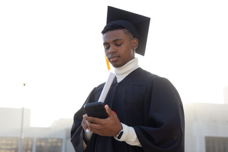 African American man standing backlit in plaza, wearing cap,gown, holding diploma, checking phone....