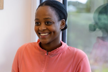 African American woman in twenties smiling, facing window at home in pink zip-up. Portrait,...