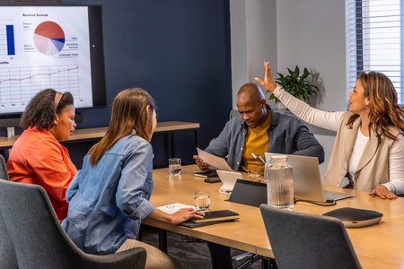 Diverse coworkers wearing blazers discussing on wooden meeting table with laptops, charts, pitcher....