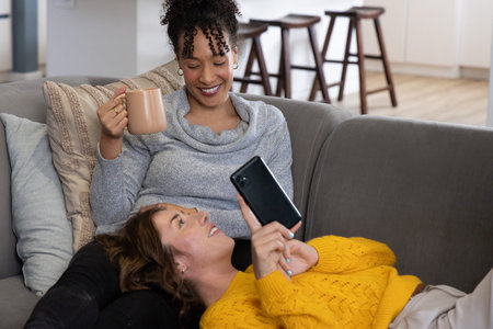 Two women reclining on gray sofa in living room, wearing knit sweaters, holding mug and smartphone. Duo, home, interior, cozy, calm, neutral, lifestyleの写真素材