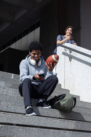 African American and Asian teenage males sitting on campus concrete steps holding football, phones....