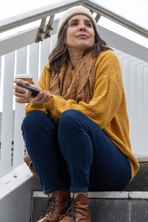 Adult woman sitting on concrete steps in beanie sweater holding paper cup and checking smartphone....