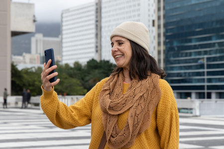 Middle-aged female standing on rooftop plaza with striped paving holding smartphone, wearing beanie....