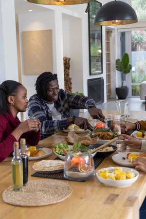 African American friends passing roasted bird around wooden dining table at modern home. Gathering,...