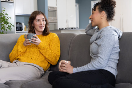 Diverse women sitting on gray sofa at home holding mugs, in yellow sweater and gray hoodie. Friends,...
