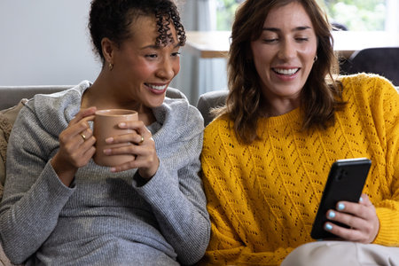 Diverse female friends leaning together, sharing smartphone on sofa in living room with beige mug....