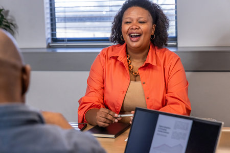 Coworkers watching woman wearing orange shirt laughing, holding pen at meeting table with laptop....