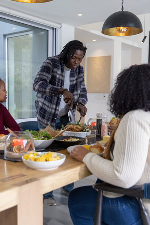 African American family sitting at dining table, man in plaid shirt carving roasted bird with knife. Gathering, home, warmth, modern, kitchen, sharing, feastの写真素材