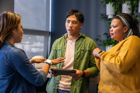 Diverse team discussing in office, denim jacket, mustard sweater, tablet and smartwatches visible....