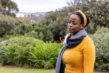 African American woman standing in park wearing mustard-yellow sweater and gray scarf, copy space....