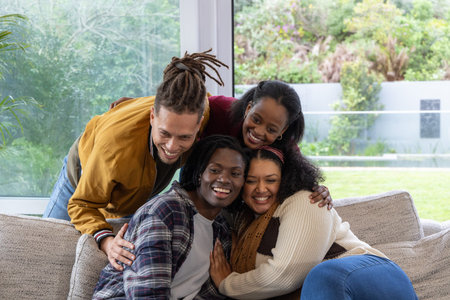 Diverse friends sitting on sofa in living room by floor-to-ceiling windows, wearing knit sweaters....