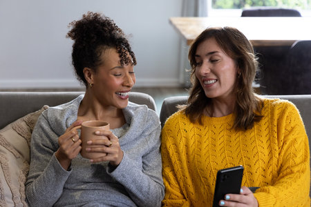 Diverse female friends sitting on gray upholstered couch in living room, holding mug and smartphone....