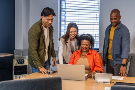 Diverse coworkers collaborating around laptop in conference room, reviewing charts, smartphone....