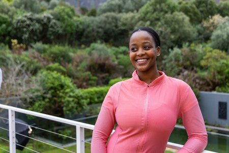 African American woman leaning on white metal balcony railing with cables, in pink zip-up jacket....