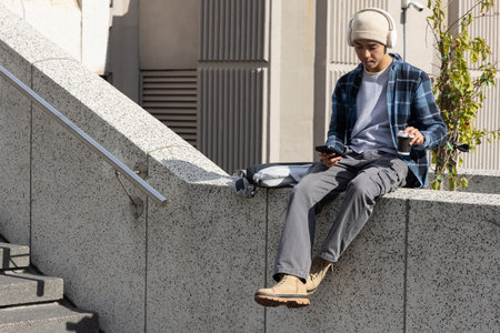 Asian man in 20s sitting in plaza, wearing headphones, holding phone and coffee, copy space. Urban,...