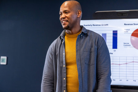 African American man presenting revenue charts on monitor in meeting room in mustard sweater....