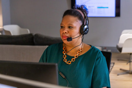 African American woman wearing headset and teal blouse sitting at desk in office using computer.