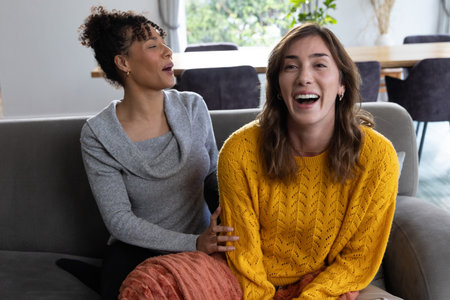 Female friends laughing, talking on grey sofa at home with mustard knit sweater, orange throw....