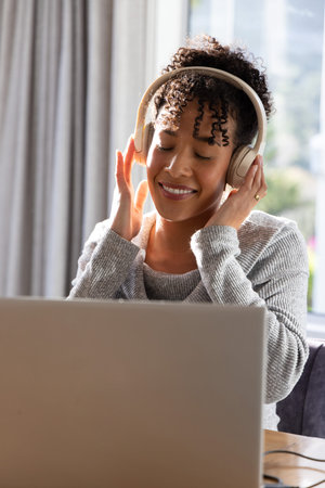 African American woman sitting at table wearing light gray sweater, holding headphones and laptop....