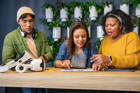 Diverse coworkers collaborating on VR headset and tablet at modern office with living wall....