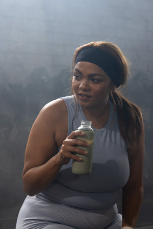 African American woman pausing workout sipping green shake from bottle in gym in sleeveless top....