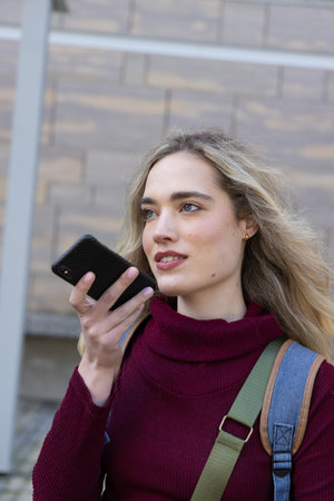 Woman in burgundy sweater holding smartphone to mouth speaking outside building carrying backpack....