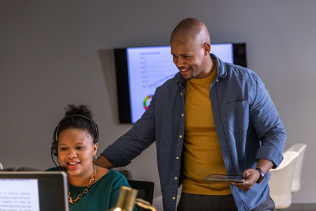 African American coworkers collaborating at office workstation, using headset and tablet by monitor....