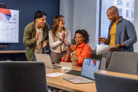 Diverse coworkers applauding seated woman wearing orange blouse at meeting room with laptops tablet....
