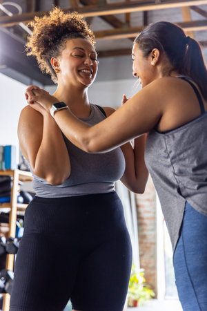Diverse female friends smiling and holding forearms while training in gym with dumbbell rack....