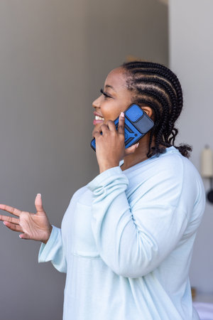 African American woman smiling and speaking on blue smartphone wearing light blue top at home....