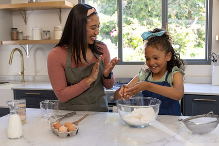 Mother and daughter wearing aprons mixing batter at home kitchen island with mixing bowl and eggs....