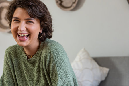 Woman sitting on sofa at home laughing wearing green knit sweater hoop earrings short necklace....