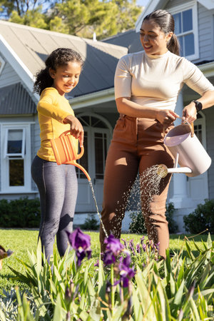 Mother and school-age daughter watering purple iris in yard using orange and white watering cans....