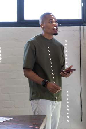 African American adult male standing by high window in bright room, holding smartphone and earbud....