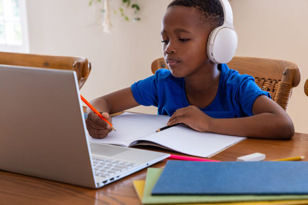 African American male child writing in spiral notebook at home table using laptop and orange pencil....