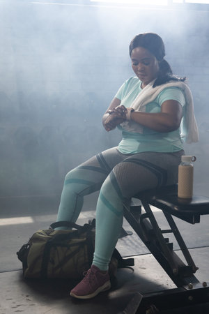 African American woman sitting on flat weight bench at gym checking smartwatch, wearing mint top....