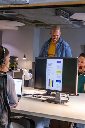 Diverse coworkers collaborating around desktop monitor at open-plan office with headsets, laptop....