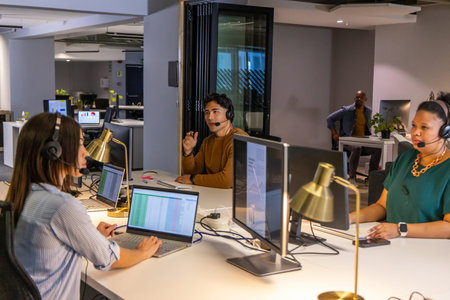 Diverse call center coworkers working in open-plan office using headsets laptops gold desk lamps....
