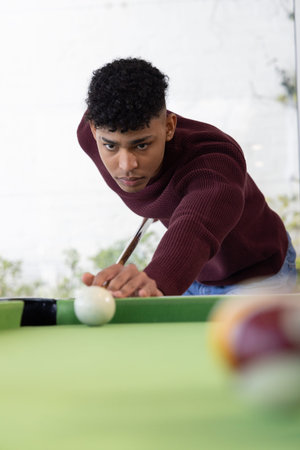 African American man leaning over pool table near window, lining up white cue ball, maroon sweater....