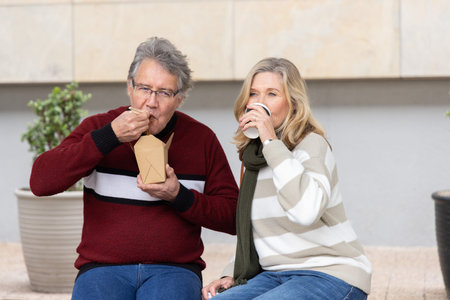 Senior couple sitting on bench in urban courtyard eating from takeout container and sipping coffee....