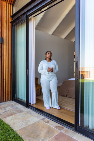 African American woman standing in sliding door holding ceramic mug looking out in pajamas. Bedroom,...