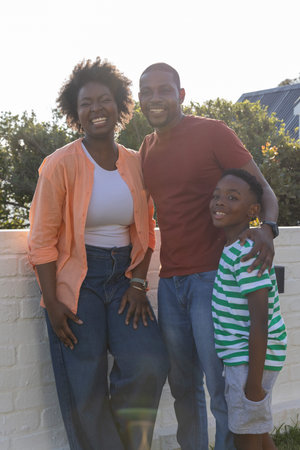 African American family with son standing smiling on patio by white brick wall, man wearing watch....
