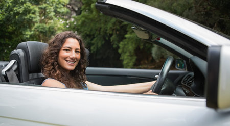 Woman wearing blue top sitting in silver convertible top down in park, reaching wheel, smiling. Car,...