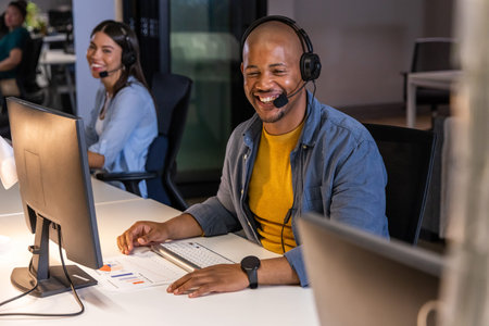 Diverse coworkers smiling and collaborating at modern office desks with headsets, monitors, charts....