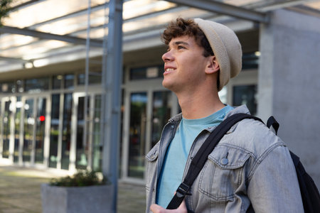 Teenage male standing at campus entrance wearing beige beanie and denim jacket, carrying backpack....