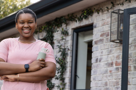 African American adult woman standing smiling on porch in pink tee holding water bottle, copy space. Brick, vines, sunlit, exterior, greenery, female, lifestyleの写真素材