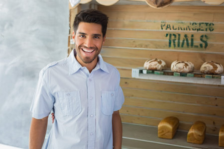 Man in his 20s standing smiling at camera in bakery stall with artisan loaves on shelves. Bread,...