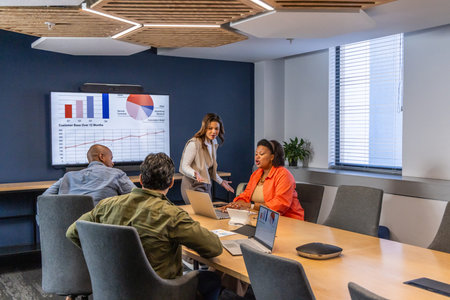 Diverse coworkers presenting charts on wall display in meeting room in office attire using laptops. Boardroom, conference, collaboration, graphs, analytics, strategy, daylight