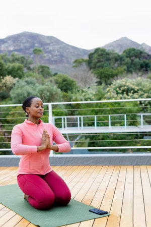 African American woman kneeling, meditating on green mat on deck in pink gear, smartphone nearby. Yoga, mindfulness, breathing, outdoor, terrace, nature, wellness