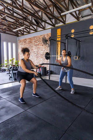 Diverse female workout partners wearing athletic clothing using battle ropes on rubber mat at gym. Rope, teamwork, training, strength, studio, focus, energy